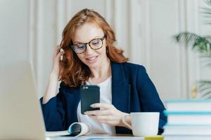 foto di una libera professionista usa lo smartphone per la comunicazione online, ha i capelli ricci rossi, indossa occhiali da vista e un costume formale, fa una pausa caffè dopo aver lavorato al computer portatile, usa il wifi gratuito