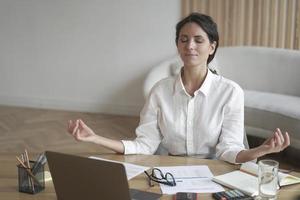 calma donna italiana con gli occhi chiusi che medita sul posto di lavoro mentre si siede al tavolo con il laptop foto