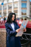 giovane donna in piedi sul lungomare della grande città e guardando la guida, turista in cerca di attrazioni. un'affascinante donna premurosa vestita alla moda con lunghi capelli scuri viaggia attraverso l'Europa foto