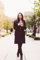 bella ragazza bruna seria intelligente che tiene una tazza di caffè in mano va a piedi per la strada di st. pietroburgo nel centro della città. affascinante donna premurosa con lunghi capelli scuri vaga da sola foto