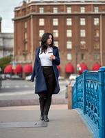 bella ragazza bruna seria intelligente che tiene una tazza di caffè in mano va a piedi per la strada di st. Pietroburgo nel centro della città sul ponte. affascinante donna premurosa con lunghi capelli scuri vaga da sola foto