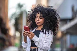 giovane donna nera capelli ricci che cammina utilizzando il telefono cellulare. sms per strada. grande città. foto