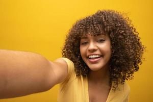 primo piano di una giovane ragazza afroamericana eccitata in maglione di pelliccia in posa isolata su sfondo giallo arancione in studio. concetto di stile di vita delle persone. simulare lo spazio di copia. facendo selfie sul cellulare. foto