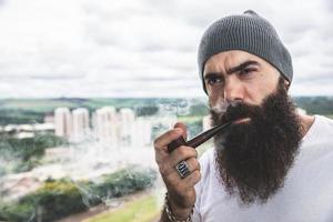 elegante uomo barbuto che fuma la pipa guardando l'orizzonte dalla cima di un edificio alto. foto