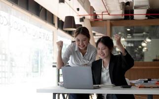 due giovani imprenditrici asiatiche mostrano gioiosa espressione di successo sul lavoro sorridendo felicemente con un computer portatile in un ufficio moderno. foto