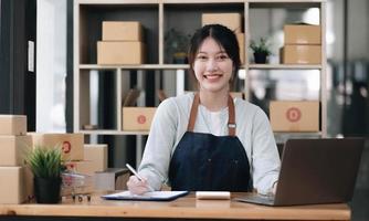 un ritratto di una giovane donna asiatica dipendente di e-commerce seduto in ufficio pieno di pacchi sullo sfondo scrivere una nota di ordini e una calcolatrice, per le piccole imprese di e-commerce e consegna. foto