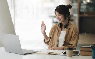 donna asiatica che indossa le cuffie sul collo usando il laptop e salutando gli amici durante la videochiamata a casa foto