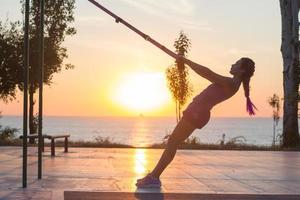 bella donna in forma in abbigliamento sportivo rosa e viola allenamento in palestra all'aperto al mattino, esercizi con cinghie di sospensione nel parco foto