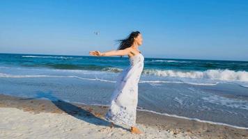 giovane bella donna vestita con un abito bianco cammina a piedi nudi sulla spiaggia estiva foto