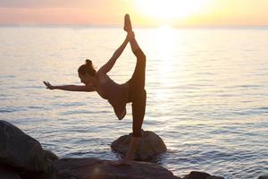 fitness razza mista donna asiatica in posa yoga sulla spiaggia del mattino, bella donna in forma pratica fitness exrxise pietre, mare mattutino o sfondo oceano foto