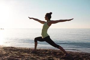fitness razza mista donna asiatica in posa yoga sulla spiaggia del mattino, bella donna in forma pratica fitness exrxise pietre, mare mattutino o sfondo oceano foto
