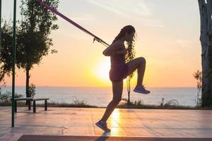 bella donna in forma in abbigliamento sportivo rosa e viola allenamento in palestra all'aperto al mattino, esercizi con cinghie di sospensione nel parco foto