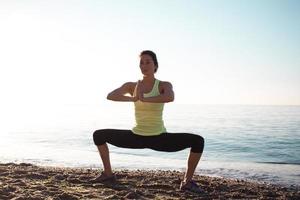 fitness razza mista donna asiatica in posa yoga sulla spiaggia del mattino, bella donna in forma pratica fitness exrxise pietre, mare mattutino o sfondo oceano foto