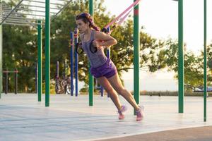 bella donna in forma in abbigliamento sportivo rosa e viola allenamento in palestra all'aperto al mattino, esercizi con cinghie di sospensione nel parco foto