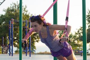 bella donna in forma in abbigliamento sportivo rosa e viola allenamento in palestra all'aperto al mattino, esercizi con cinghie di sospensione nel parco foto