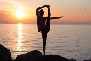 fitness razza mista donna asiatica in posa yoga sulla spiaggia del mattino, bella donna in forma pratica fitness exrxise pietre, mare mattutino o sfondo oceano foto