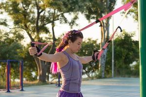 bella donna in forma in abbigliamento sportivo rosa e viola allenamento in palestra all'aperto al mattino, esercizi con cinghie di sospensione nel parco
