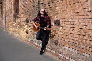 giovane maschio con capelli lunghi e tatuaggio suona la chitarra acustica all'aperto per strada foto