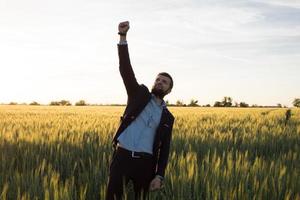 concetto di vincitore, felice giovane imprenditore salta con le mani alzate nei campi, piante di grano estivo foto