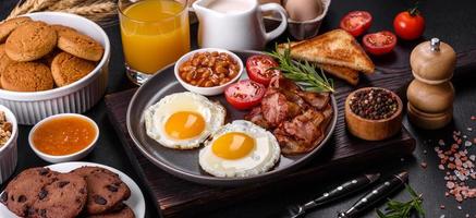 una deliziosa colazione sana e nutriente con uova fritte, pancetta, fagioli, un bicchiere di succo, biscotti d'avena, latte e marmellata foto