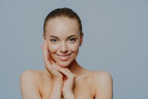 studio di una bella donna sorridente che tocca il viso e gode di una pelle impeccabile, si trova a spalle nude, ha una grande fronte, posa su sfondo blu, applica cosmetici per la cura della pelle. tenerezza, trucco foto