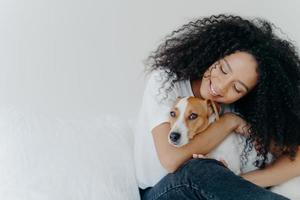 foto di donna attraente con acconciatura afro riccia, coccole e animali domestici cane con sorriso, esprime amore, gode di un'atmosfera domestica accogliente, posa su sfondo bianco con spazio vuoto per la promozione