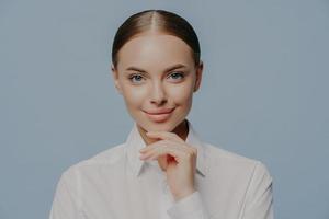 La donna d'affari attraente e sicura di sé tiene il mento e guarda direttamente la fotocamera, indossa un'elegante camicia bianca, ha una pelle sana e un trucco naturale, isolata su sfondo blu. donne, affari, carriera foto