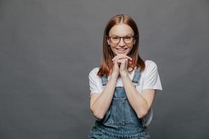 l'inquadratura orizzontale di una donna felice e contenta tiene entrambe le mani sotto il mento, ha un sorriso sul viso, vestita con una tuta di jeans, si trova su uno sfondo grigio con spazio per la copia per il tuo contenuto promozionale. foto