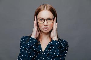 headshot di una seria insegnante femminile tiene le mani sulla testa, ha un'espressione facciale sicura di sé, indossa occhiali, camicia bianca e nera a pois, isolata su sfondo grigio. tiro orizzontale foto