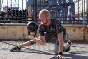 giovane atleta maschio barbuto che si allena nella zona industriale in una giornata di sole, esercizi di kettlebell all'aperto, fondo urbano foto