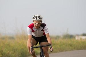 ciclista in casco e abbigliamento sportivo che si allena da solo su strada di campagna vuota, campi e alberi sullo sfondo foto