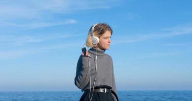 la giovane donna bella ascolta la musica con le cuffie all'aperto sulla spiaggia contro il cielo blu soleggiato foto