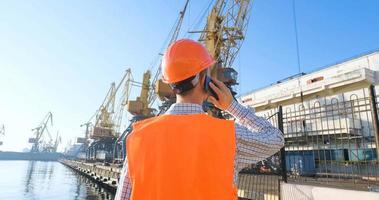 lavoratore maschio del porto marittimo con casco arancione e sicurezza ovest, gru e sfondo del mare foto