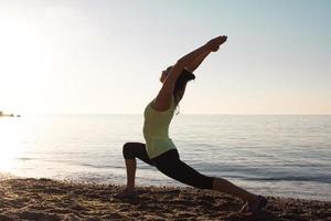 fitness razza mista donna asiatica in posa yoga sulla spiaggia del mattino, bella donna in forma pratica fitness exrxise pietre, mare mattutino o sfondo oceano foto