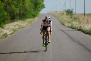 ciclista in casco e abbigliamento sportivo che si allena da solo su strada di campagna vuota, campi e alberi sullo sfondo foto