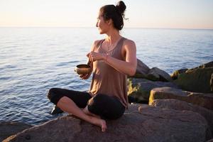 la giovane donna sportiva gioca sulla ciotola tibetana e si siede sulle pietre, bella donna asiatica che guarda l'alba nel mare nella posa di yoga foto