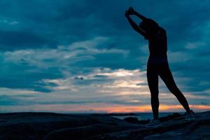 silhouette donna allenamento al mattino sulla spiaggia di pietra con bellissimo cielo all'alba. donna in forma che allunga il corpo prima dell'allenamento. esercizio per uno stile di vita sano. allenamento fuori porta. paesaggio naturale. libertà foto