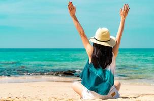 felice giovane donna in stile casual moda e cappello di paglia sedersi sulla spiaggia di sabbia. rilassati e goditi le vacanze sulla spiaggia del paradiso tropicale con acqua verde smeraldo. ragazza in vacanza estiva. vibrazioni estive. foto