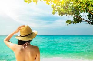 il punto di vista posteriore della giovane donna asiatica sexy e felice indossa il cappello di paglia che si rilassa e si gode la vacanza sulla spiaggia del paradiso tropicale. ragazza in piedi sulla spiaggia e guardando il mare durante le vacanze estive. vibrazioni estive. foto