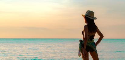 il punto di vista posteriore della giovane donna asiatica felice in costume da bagno nero e cappello di paglia si rilassa e si gode la vacanza sulla spiaggia del paradiso tropicale al tramonto. ragazza alla moda per le vacanze estive. modella sexy di bellezza. stile di vita elegante foto