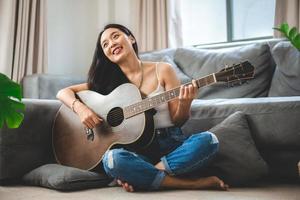 donna asiatica che suona musica con la chitarra a casa, giovane donna chitarrista musicista stile di vita con strumento di arte acustica seduto per suonare e cantare una canzone che fa suono in hobby nella stanza di casa foto