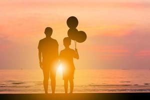 silhouette di madre e figlio in piedi a guardare il tramonto sulla spiaggia con palloncino in mano. foto