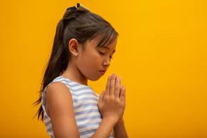 bambina che prega su sfondo giallo. piccola ragazza asiatica che prega a mano, mani giunte nel concetto di preghiera per fede, spiritualità e religione. foto