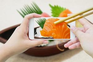 le bacchette tengono il sashimi di salmone nel telefono cellulare foto