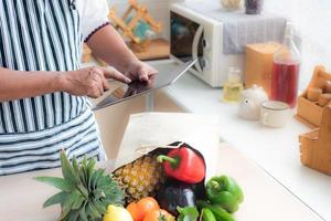 primo piano della mano dello chef utilizzando tablet per acquistare frutta e verdura online. e ci sono frutta e verdura e sacchetti di carta sul tavolo della cucina. shopping online e concetto di consegna. foto