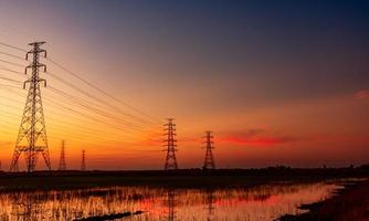 traliccio elettrico ad alta tensione e filo elettrico con cielo al tramonto. pali elettrici. concetto di potenza ed energia. torre di griglia ad alta tensione con cavo metallico in un campo di riso vicino a una fabbrica industriale. foto