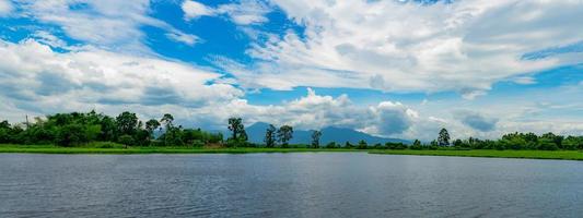 bella vista panoramica del lago di fronte alla montagna con cielo azzurro e nubi cumuliformi bianche. albero verde e campo in erba intorno allo stagno. clima tropicale in estate. paesaggio naturale. aria fresca. foto