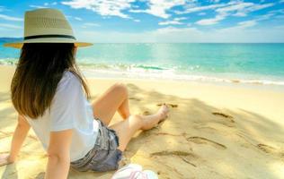 felice giovane donna in camicie bianche e pantaloncini seduti sulla spiaggia di sabbia. rilassarsi e godersi le vacanze sulla spiaggia del paradiso tropicale con cielo blu e nuvole. ragazza in vacanza estiva. vibrazioni estive. giorno felice. foto