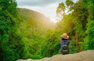 la donna felice viaggia da sola nei boschi. la donna attiva di buon umore si siede sulla pietra nella valle verde con l'albero denso nella foresta. felici vacanze. vista posteriore di donna felice indossare zaino, smart band e cappello. foto