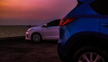 primo piano suv blu di lusso e auto bianca parcheggiata su un'area di parcheggio in cemento accanto alla spiaggia la sera con un bellissimo cielo viola al tramonto. viaggio su strada in vacanza estiva sulla spiaggia del mare tropicale. foto